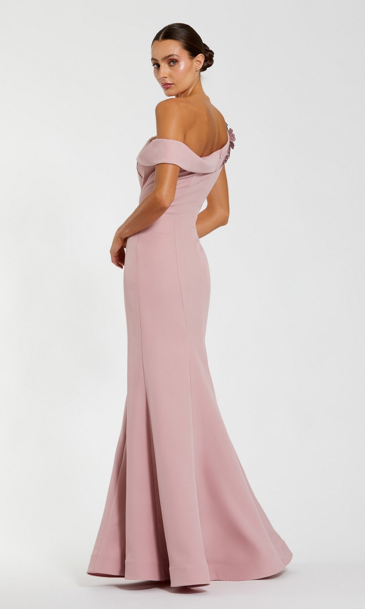 A woman models the Mac Duggal 11947 long formal dress, a blush pink off-the-shoulder crepe gown with a subtle mermaid silhouette, standing and gazing over her shoulder against a plain background.